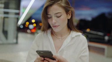 Kulaklıklı odaklanmış genç bir kadın, gece boyunca iyi aydınlatılmış bir metro istasyonunda akıllı telefon kullanıyor. Arka planda hareket eden araçlar ve şehir ışıkları görülüyor.
