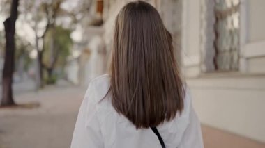 Güneşli bir caddede yürürken dalgalı saçları olan genç bir kadın neşe ve güven dolu bir şekilde gülümsüyor. Genç Kadın Güneşli Günlerin Tadını Çıkartıyor