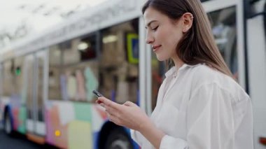 Beyaz bluzlu gülümseyen genç bir kadın renkli bir otobüsün yanında dururken akıllı telefonunu kullanır. Şehir yaşamını ve bağlantılarını kaydetmek. Şehir Otobüsü Yakınındaki Akıllı Telefon Kullanan Genç Kadın