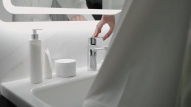 Çağdaş bir banyoda, cilt ürünleriyle çevrili musluğu açan ellerin yakın çekimi. Temiz ve minimalist tasarım hijyen ve kendine bakmanın altını çiziyor. Banyodaki Musluğu Açan Eller