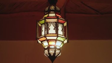 Tavandan sarkan renkli cam panellerle süslü Fas lambası. Sıcak ışık, karmaşık metal işçiliğini aydınlatıyor. Geleneksel tasarım, kültürel estetik ve sanatsal işçilik. İç kısımlar.