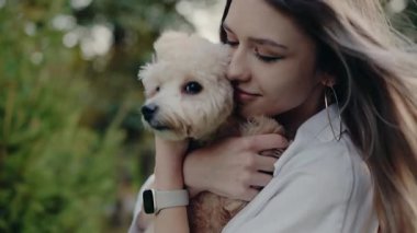 Genç bir kadın küçük tüylü köpeğini nazikçe bir park ortamında tutuyor. Sahne, sevgi, ilgi ve arkadaşlığı çağrıştırıyor. Kadın ve evcil hayvanı açık havada hassas bir anı paylaşıyorlar..