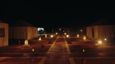 Fas 'ta bir kamp alanında, geleneksel çadırlar arasındaki yolu aydınlatan parlak fenerlerle dolu sakin bir çöl sahnesi. Huzurlu ve davetkar bir macera ve sükunet ortamı..