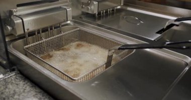Bir restoranın mutfağında kızartma kızartıcısının yakın çekimi. Kızartmalar kızgın yağ içinde kaynarken maşalar metal sepeti kaldırıyor. Paslanmaz çelik fritöz ışığı yansıtır..