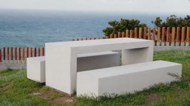 Somut Okyanus manzaralı Piknik Masası. Yeşil çimenlerin arasına kurulmuş modern beton piknik masası huzurlu bir sahil bölgesinde çarpıcı bir okyanus manzarası sunuyor..
