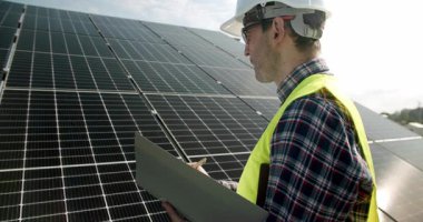 Erkek mühendis güneş panellerini inceliyor. Üniformalı bir yetişkin fotovoltaik panelleri kontrol ediyor ve güneş enerjisi istasyonuyla ilgili çalışırken günlüğe not alıyor.