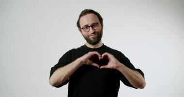 İnsan elleri ile kalbi şekillendirir ve kamerayı gösterir. Gülümsüyor ve kameraya bakıyor. Siyah bir tişört ve gözlük takıyor. Sakalı var ve saçları arkadan bağlanmış..