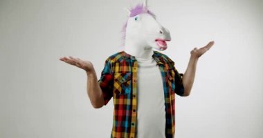 Tek boynuzlu at maskesi ve renkli ekose gömlek giyen bir adam beyaz arka planda duruyor. Omuzlarını silker ve elleriyle hareket eder, sanki ne olduğunu sorar gibi. Video, mizah ve kaprisleri çağrıştırıyor..