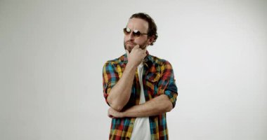 Renkli tişörtlü ve güneş gözlüklü düşünceli bir adam beyaz bir arkaplana kafa yoruyor. Çenesini tutuyor, düşüncelerde kayboluyor, bir karar ya da fikir üzerinde kafa yoruyor..