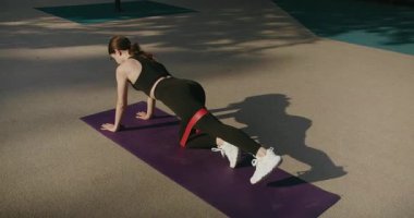 Spor giyinen esmer kadın sporcu, direniş bandıyla tahtada kal ve bacağını kaldır. Sportif kadın dışarıda egzersiz minderinde çalışıyor. Yoga yapan bir kadın yoga minderinde poz veriyor..