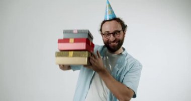 Gözlüklü ve parti şapkalı mutlu bir adam bir yığın hediyeyi dengeliyor. Gülümser ve kutuları gösterirken ayarlar. Beyaz arka planda renkli hediyeler.