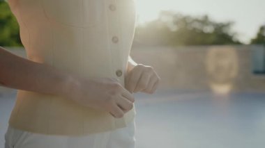 Açık havada açık havada, yeşil arkaplanı bulanık bir şekilde bej yeleğini ilikleyen bir kadının yakın çekimi. Sakinlik ve hazırlığın büyüleyici görüntüsü. Kadın Düğmeli Bej Yelek