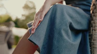 Kot bir elbisenin üzerinde nazikçe dinlenen bir kadının eli. Arka plan, doğal ışık alan bir açık hava ortamını gösteriyor. Görüntü, dinginlik ve kadınlığı çağrıştırıyor..