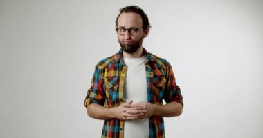 Gözlüklü ve ekose gömlekli sakallı bir adam beyaz bir arka plana karşı duruyor, hoşnutsuz görünüyor ve ellerini karnının önünde sıkıca tutuyor. Düşünüyor veya bekliyor gibi görünüyor..