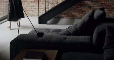 Uzun siyah elbiseli bir kadın odada yürüyordu. Köşedeki kanepe ve kitaplığı olan lüks modern ev iç mimarisi. Moda mobilyalar. Minimalist İç İşleri. Lüks Zarif Oda