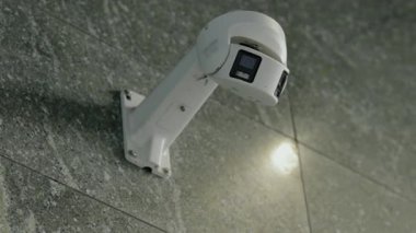 Güvenlik kamerası fayanslı bir duvara monte edilmiş. Kamera beyazdır ve beyaz bir tepeye bağlıdır. Duvarda küçük bir ışık görünüyor. Bu izleme ve güvenlik sağlar. Modern Güvenlik Kamerası