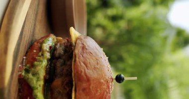 Peynirli, sebzeli ve soslu sulu sığır etli burger bulanık arka planda masada yavaşça döner. Biftek burgeri bir restoranın terasında servis edilir. Hikayeler için dikey video.
