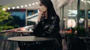 Siyah deri ceketli düşünceli bir kadın akşamları çatı katındaki bir terasta tek başına oturuyor. Etrafı tel ışıklarla çevrili. Siyah Deri Ceketli Dalgın Kadın Çatıdaki Kafe Terasında Oturuyor
