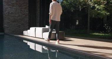 Şık bir genç adam modern bir yaşam tarzını ve lüks bir konutta yaşamayı yansıtan çağdaş bir kanepe seti ve yemyeşil bir havuzun yanında rahatça yürüyor. Modern Ev Harici