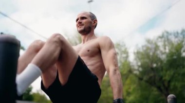 Kaslı bir adam, şehir parkı ortamında paralel barlara daldırır. Üstsüz ve antrenmanına odaklanmış durumda. Bu video fitness, kuvvet eğitimi ve sağlıklı yaşam tarzını temsil ediyor..