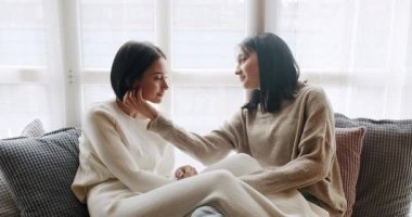 Low angle handheld shot of young lesbian couple smiling and touching hands gently while sitting on sofa against window in light living room at home. Lifestyle and relationships. LGBT concept