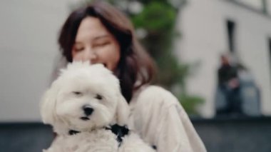 Genç kadın küçük beyaz köpeğini kollarında tutuyor ve gülümsüyor. Şehir caddesinde dışarıdalar. Köpeğin siyah bir yakası var. Kadının uzun kahverengi saçları var ve açık bej bir gömlek giyiyor..