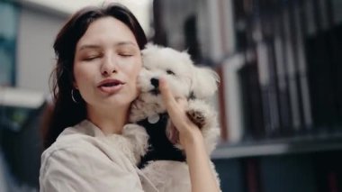 Genç kadın küçük beyaz köpeğini sevgiyle tutuyor. Açık havada, evcil hayvan ve sahibi arasındaki bağı gösteren hassas bir anı paylaşıyorlar. Evcil hayvan projeleri için mükemmel. Sevgi, ilgi, arkadaşlık.