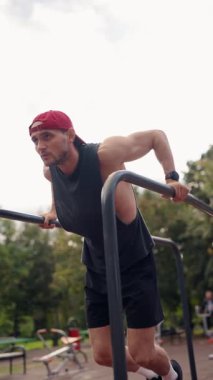 Kararlı bir erkek sporcu, açık hava spor parkındaki paralel barlara dalış yapar, dayanıklılık, zindelik ve egzersize adanmışlık gösterir. Dikey video, odaklanırken erkeğin çabasını yakalar