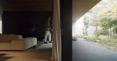 Profesyonel bir adam şık, modern bir evin engin cam duvarının ardındaki huzurlu bahçe manzarasını seyrediyor. Modern Ev Cam Duvarından Doğayı Düşünen Adam