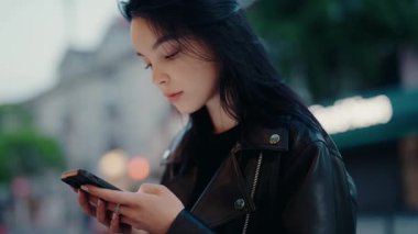 Alacakaranlık 'ta şehir merkezinde akıllı telefon kullanan siyah deri ceketli genç bir kadın. Telefonunu kontrol ederken odaklanmış görünüyor. Genç Kadın Alacakaranlık Şehri 'nde açık havada akıllı telefon kullanıyor.