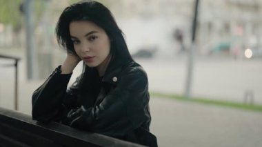 Siyah deri ceketli genç bir kadın, kentin arka planında bir bankta oturuyor, rahat ve düşünceli bir ruh hali içinde. Şık Genç Esmer Kadın Şehir Merkezinde Park Bench 'te Dinleniyor