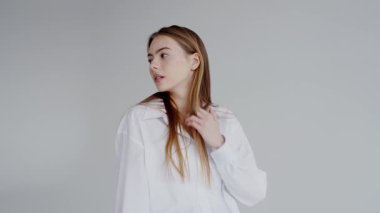 Gevşek beyaz gömlek ve açık kot pantolon giyen sakin ve şık genç bir kadın zarif bir şekilde oturur, minimalist bir ortamda kameranın önünden yavaşça bakar..