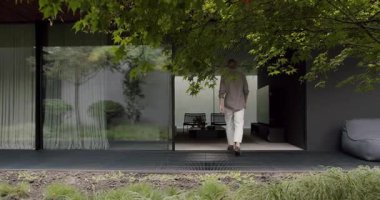 Şık giyinmiş şık bir adam yemyeşil ve zarif şeffaf perdelerle çevrili modern bir eve doğru yürüyor. Modern Ev Harici Erkek Tarz Evi 'ne Giriyor