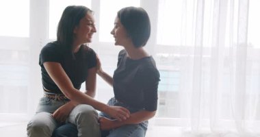 Low angle handheld shot of young lesbian couple smiling and touching hands gently while sitting on sofa against window in light living room at home. Two women sharing love and support holding hands.