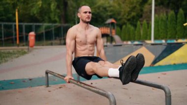 Kaslı bir adam parktaki paralel çubuklarda dibe vurur. Uygun biçime odaklanıyor ve hareketleri kontrol ediyor. Güç ve uygunluğa olan bağlılığını gösteriyor. Antrenmanın sakin atmosferi.