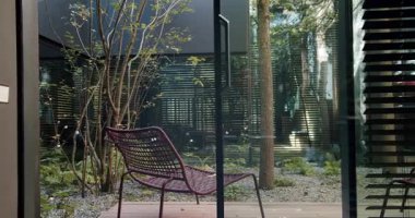 Ağaçlarla çevrili ahşap bir güvertede modern bir hasır sandalye ve çağdaş bir evin yansıtıcı camları. Modern Home Garden 'daki ahşap güvertede şık bir sandalye.