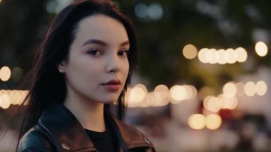 Deri ceket giyen kendine güvenen bir kadının portresi, akşamları açık havada güzel bokeh ışıkları ve sakin bir ifadeyle dikiliyor. Deri Ceketli Kadın Bokeh Işıklı Açık Hava Poz Veriyor