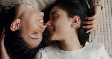 Low angle handheld shot of young lesbian couple smiling and touching hands gently while sitting on sofa against window in light living room at home. Two women sharing love and support holding hands.
