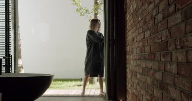 Profesyonel bir kadın şık bir ofiste, koyu renkli tuğla bir duvar ve modern mobilyaların yer aldığı geniş bir pencerenin yanında düşünceli duruyor. Brick Wall Interior 'la Modern Ofiste Duran Kadın