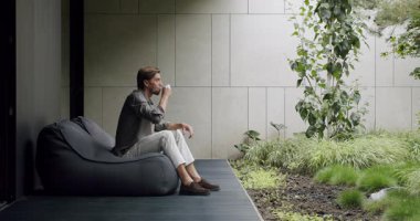 Genç bir adam şık bir evin yanındaki huzurlu, modern bir bahçede armut çuvalında otururken akıllı telefon kullanıyor. Çağdaş Bahçe Ayarlarında Telefondaki Adam, Modern Ev