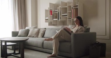 Rahat bir kanepede oturan kadın modern, rahat bir oturma odasında şık bir raf ve minimalist tasarımı olan bir kitap okuyor. Modern oturma odasında şık raflarla kitap okuyan bir kadın.
