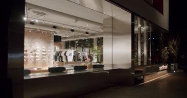 Geceleri şehir moda mağazası. Pencerede kaykay, kıyafet ve ayakkabılar sergileniyor. Tuğla ve metalle modern tasarım. Moda, havalı bir hava. Perakende, iş ve yaşam tarzı projeleri için mükemmel.