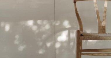 Güneş ışığı bir pencereden akar, örülmüş koltuğu olan basit, zarif, ahşap bir sandalyenin arkasındaki krem bir duvarda dans eden yaprakların gölgelerini oluşturur. Doğal ışık sıcak ve davetkar bir atmosfer yaratır.,