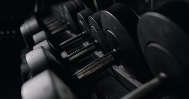 Salondaki bir askıya düzgünce yerleştirilmiş siyah halterli bir sıra halterin düşük ışık görüntüsü ortam aydınlatmasıyla şekillenen dokulara ve kontrastlara odaklanıyor. Dark Gym Ayarında Bir Rafta Ağırlıkların Görünümü