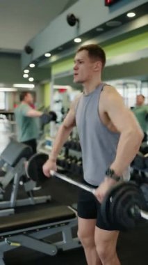 Ağırlık kaldırma seansı sırasında spor salonunda odaklanmış genç bir erkek. Güç ve zindelik gösterisi yapıyor. Çağdaş bir ortamda halter tutuyor. Ağırlık kaldıran genç adam. Dikey video.