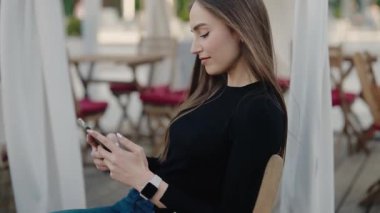 Genç bir kadın açık bir kafede oturuyor, akıllı telefonunu kullanıyor ve akıllı bir kaset takıyor. Olay yeri rahat ve rahat. Açık Cafe 'de Dinlenen Kadın Akıllı Telefon Kullanıyor ve Akıllı Seç Giyiyor