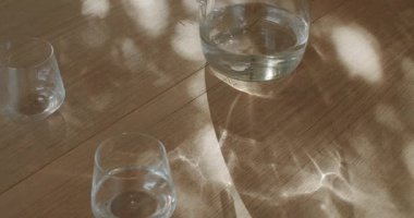 Güneş ışığı pencereden akar, ahşap bir masanın üzerinde dans eden gölgeler bırakır, cam sürahi su iki temiz bardağın yanında oturur..