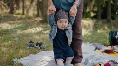 Güneşli bir günde bebeğine ilk adımlarını atmasında yardım eden bir anneyi gösteren aile pikniğinin iç açıcı bir sahnesi. Güneşli bir parkta piknik yaparken neşeli aile zamanı