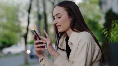 Uzun saçlı ve bej ceketli genç bir kadın, dışarıda, yeşilliklerle çevrili bir parkta otururken telefonuna odaklanıyor. Parkta Akıllı Telefon Kullanan Genç Kadın