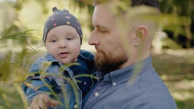 Sonbahar kıyafetleri içinde bebeğini tutan bir babanın dokunaklı sahnesi, doğal yeşil bir zemine karşı. Doğada Baba ve Bebek arasındaki hassas an.